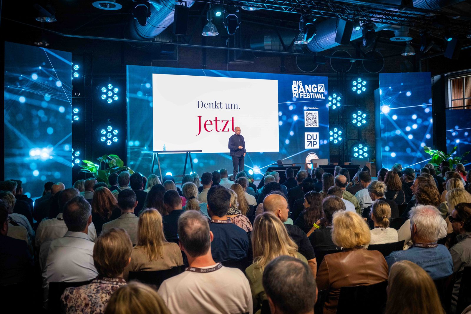Christian Steiger auf der Bühne des BIG BANG KI FESTIVALs, davor ein voller Saal, auf der Leinwand steht Denkt um. Jetzt