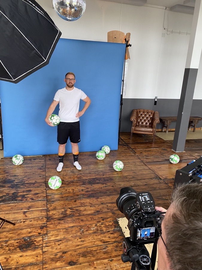 Fotoshooting vor blauem Hintergrund: ein Mann in Fußballkleidung hält einen Ball, um ihn liegen weitere Bälle, vorne eine Kamera