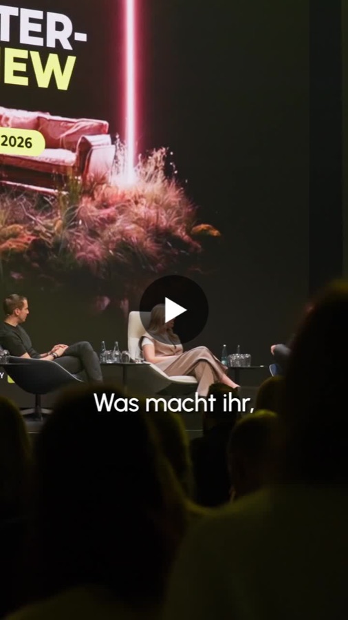 Vorschaubild zu einem Video: Bühnengespräch zweier Menschen in Sesseln vor Publikum, Untertitel „Was macht ihr,“